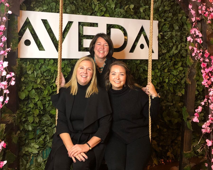 Three women smiling and posing for a photo in front of a wall with a sign that reads "Aveda".
