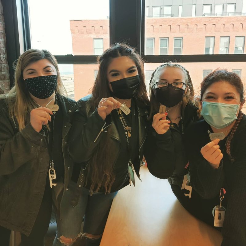 Four Institute Students wearing face masks and posing for a picture, proudly holding up their Aveda dog tags.